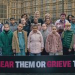 A group of children and adults standing together with a sign that says: 'Hear Them Or Grieve Them'