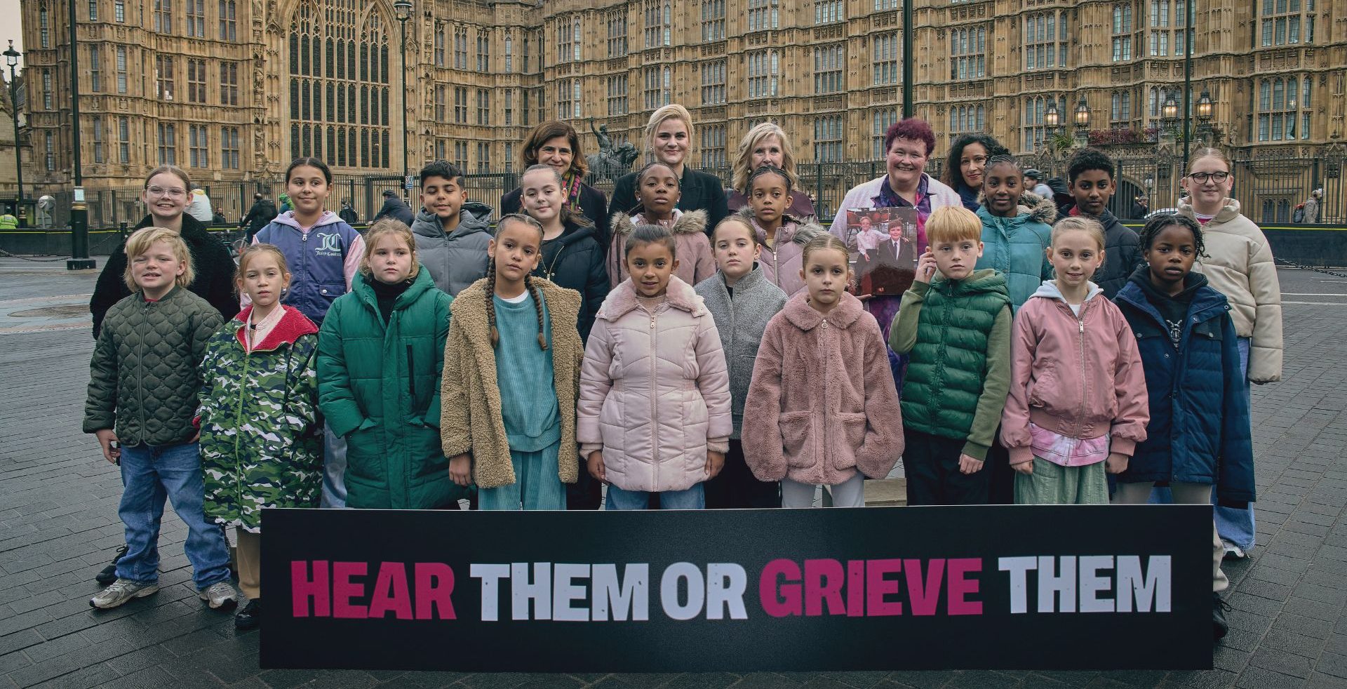 A group of children and adults standing together with a sign that says: 'Hear Them Or Grieve Them'