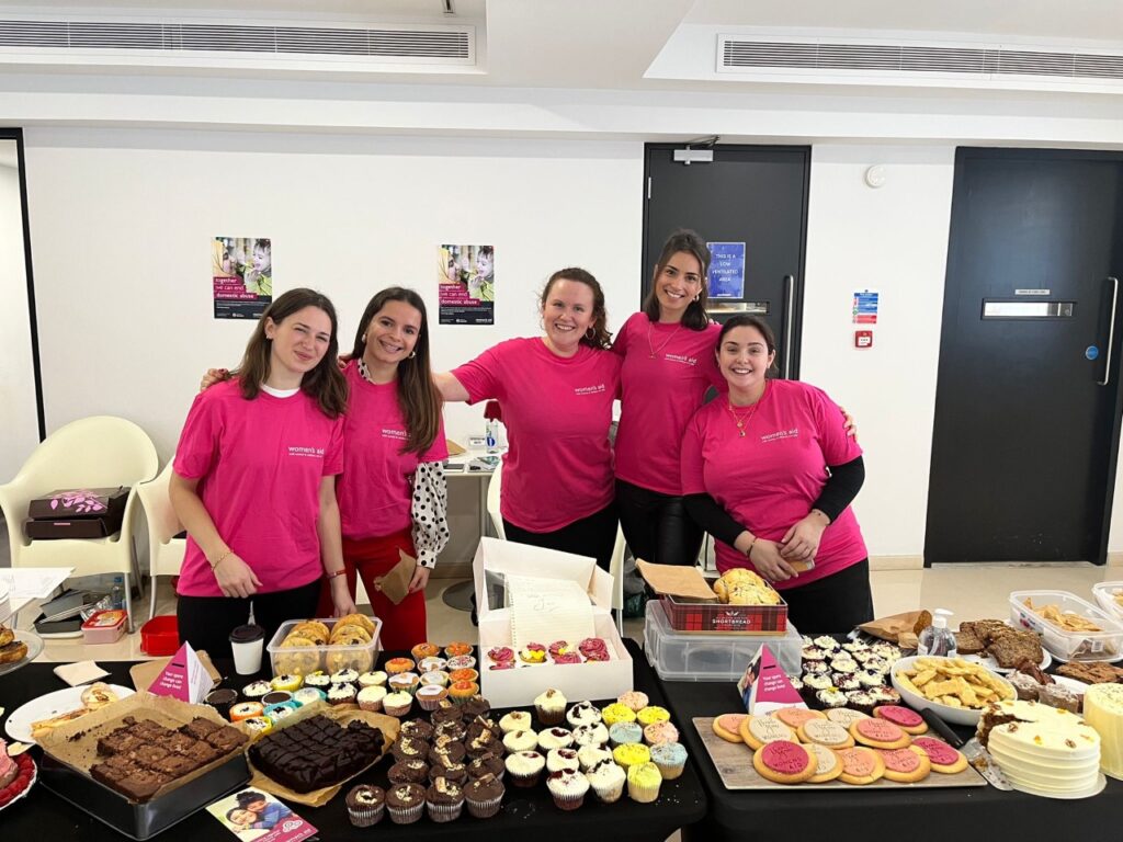 A group of women in pink Women's Aid t-shirts standing behind a table full of baked goods